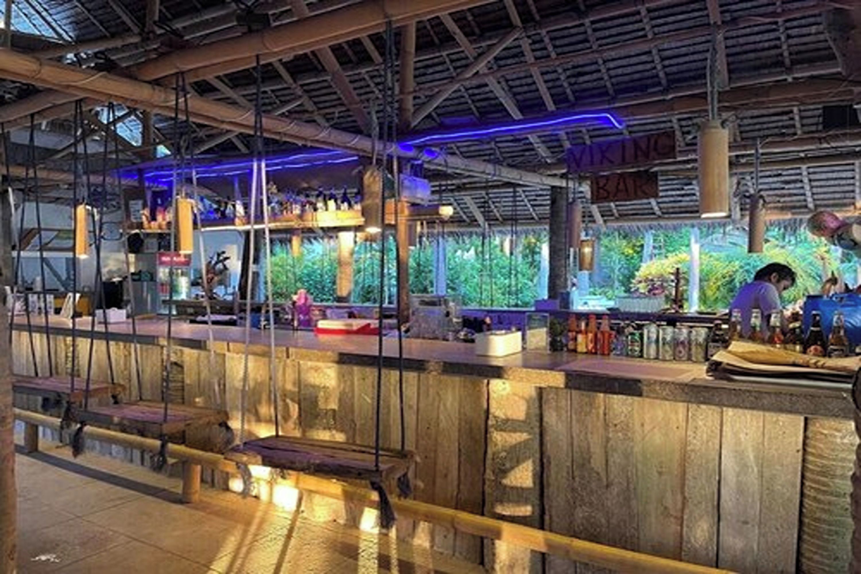 Una zona de bar con un mostrador de madera rústica, luces colgantes y vistas a la vegetación exterior.