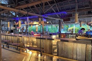 Eine Bar mit einer rustikalen Holztheke, hängenden Lampen und Blick auf Grünflächen im Freien.