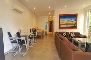A modern living room with a brown sofa, a dining area with white chairs, a painting on the wall, and a hallway leading to another room. - Pili Beach Resort Agmanic By Zuzu (Santa Fe)