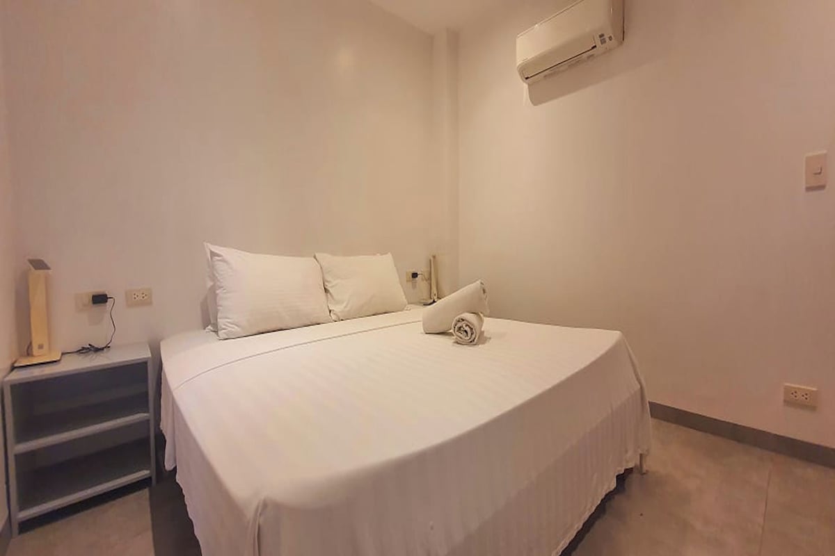 A neatly made bed with white bedding in a room with a white wall and a small bedside table.