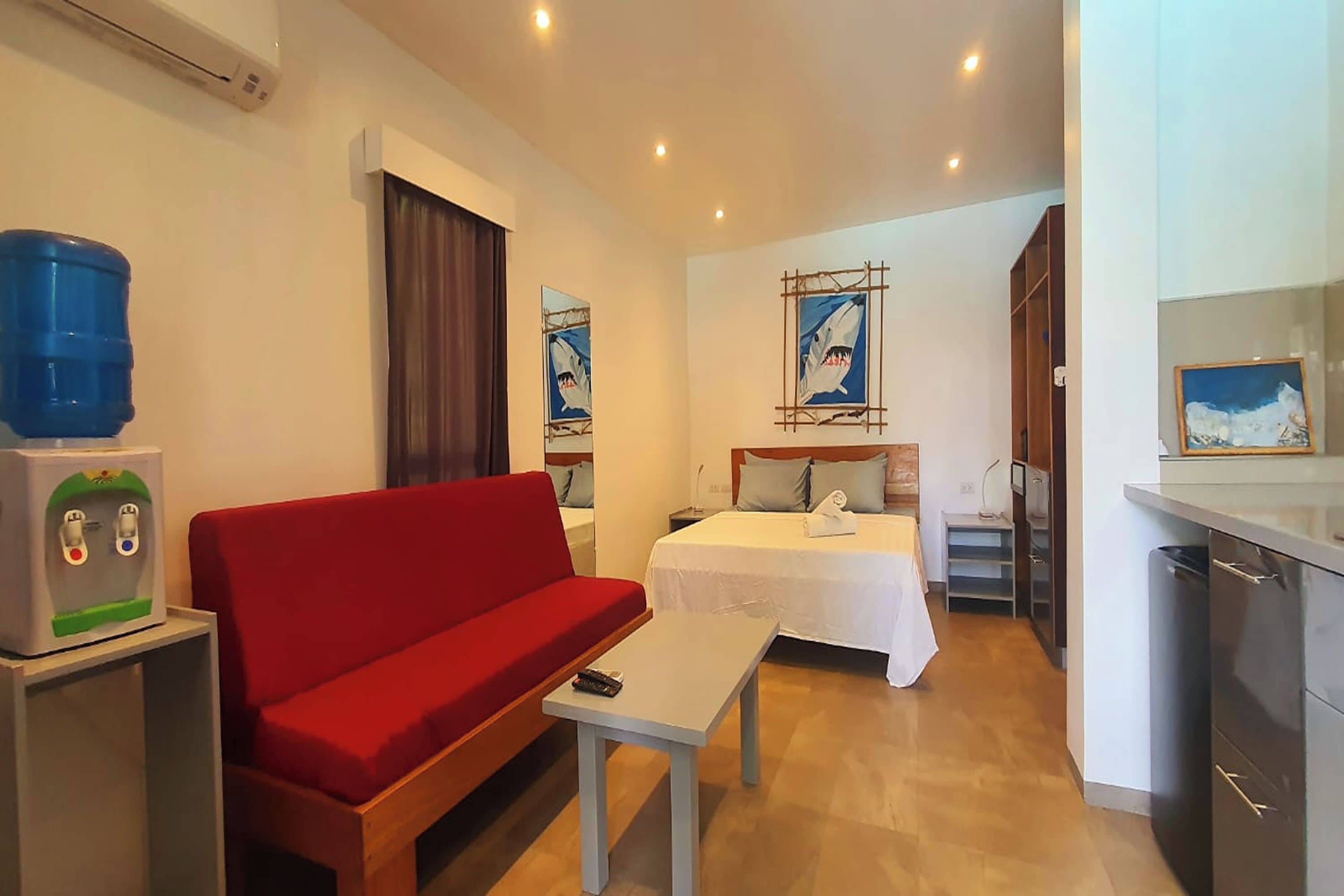 A hotel room with a red sofa, a small table, a bed with a white bedspread, a water dispenser, and a painting of a shark on the wall.