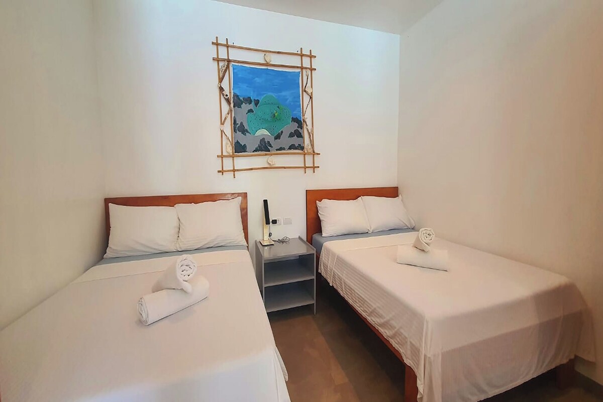 Ein Hotelzimmer mit zwei Einzelbetten, beide mit weißer Bettdecke und passenden Kissen. Dazwischen befindet sich ein kleiner Nachttisch mit einer Lampe und einer Steckdose. Über den Betten hängt ein gerahmtes Landschaftsbild.