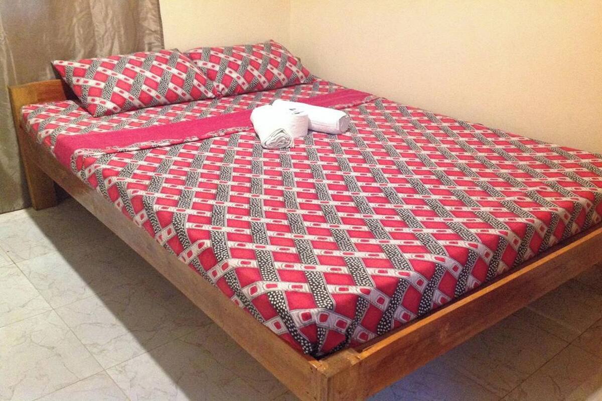 Uma cama de solteiro com colcha estampada e almofadas combinando, uma toalha branca enrolada e uma cabeceira de madeira.