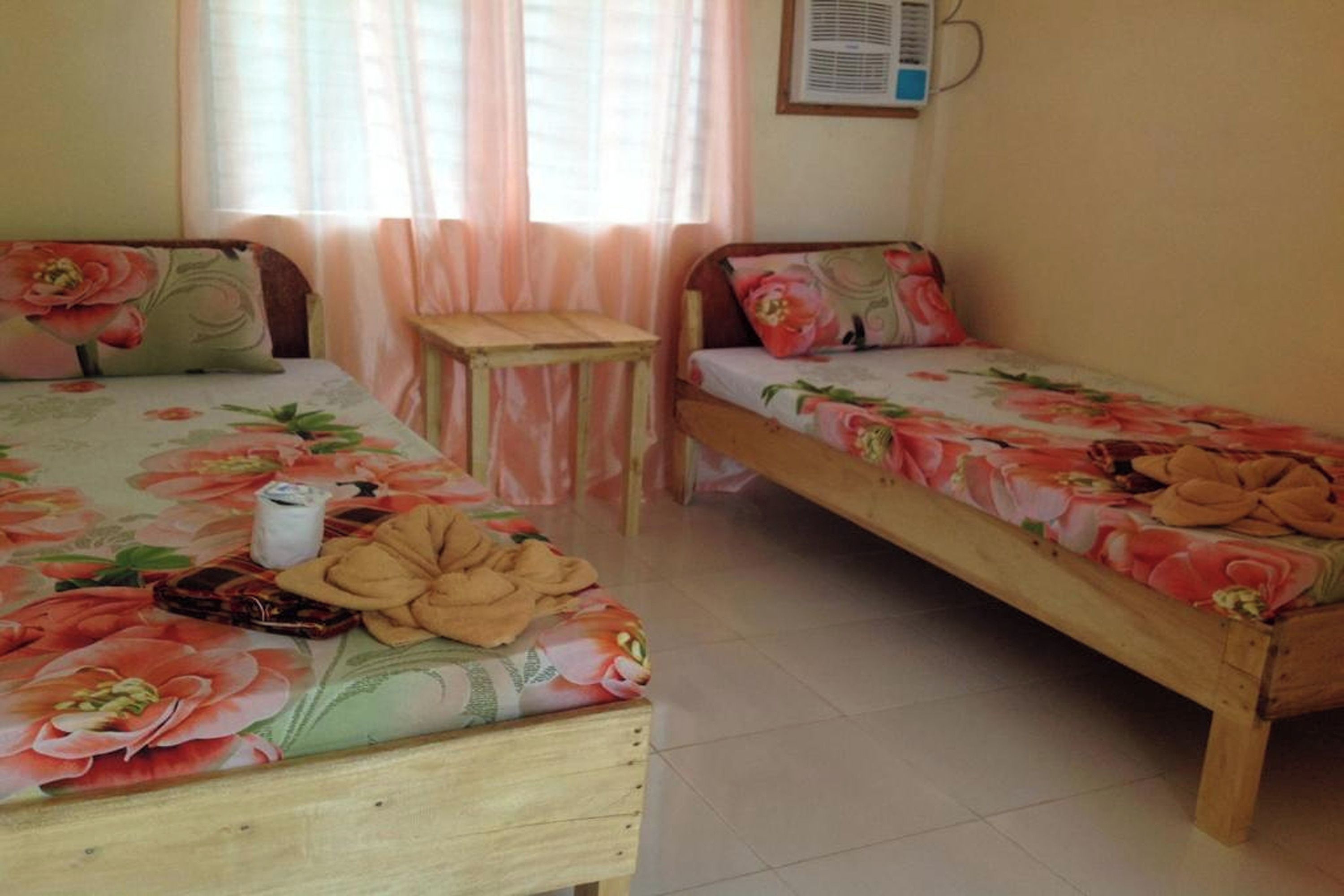 A room with two wooden beds, each with floral bedding. There is a small wooden table between the beds.
