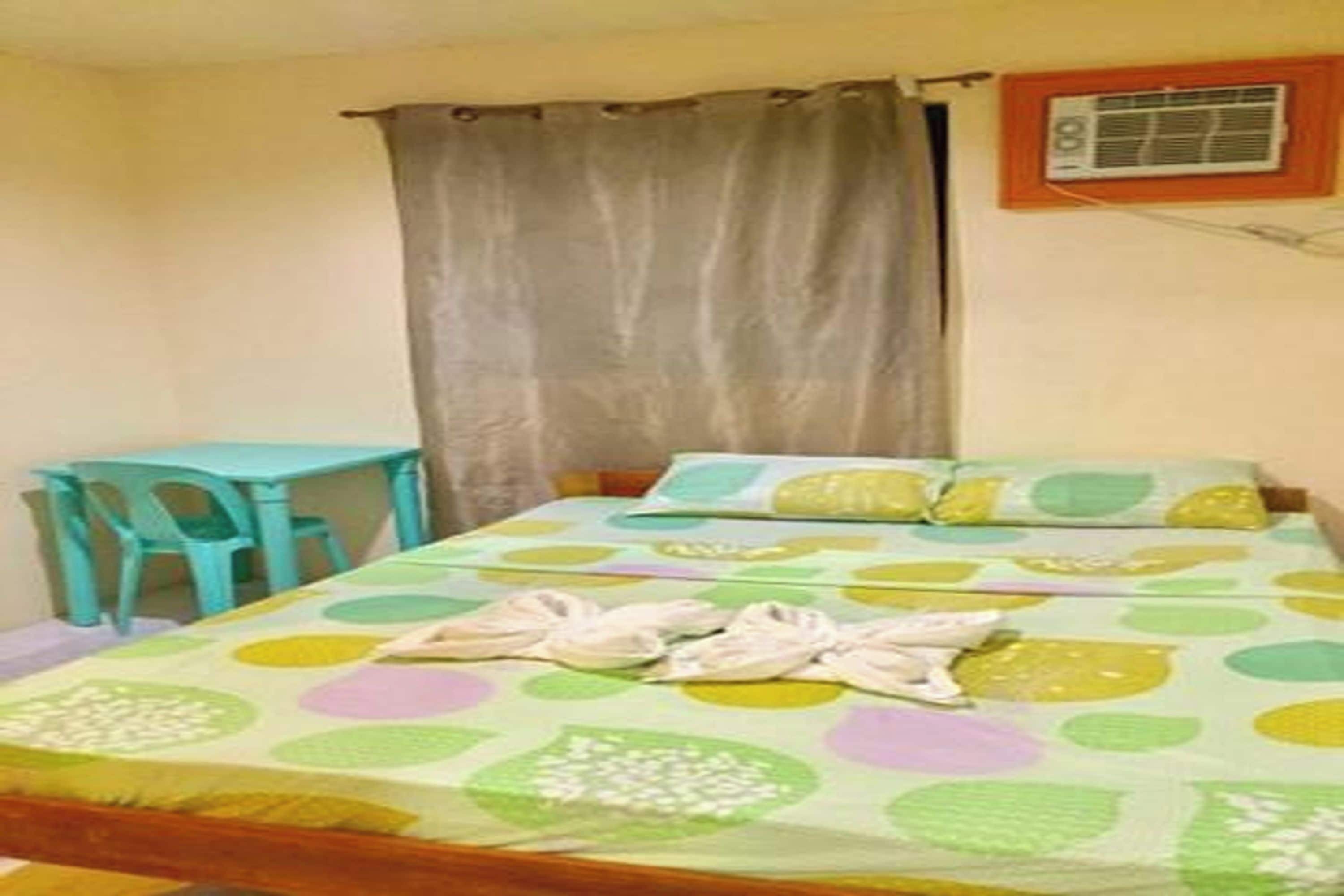 Uma cama de solteiro com um edredom estampado com bolinhas coloridas, uma mesinha e uma cortina.