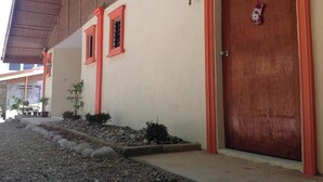 A building with a beige facade, red-framed windows, a wooden door, and a small garden area with rocks and plants. - Casa Paliton Siquijor Near Beach By Zuzu (San Juan)