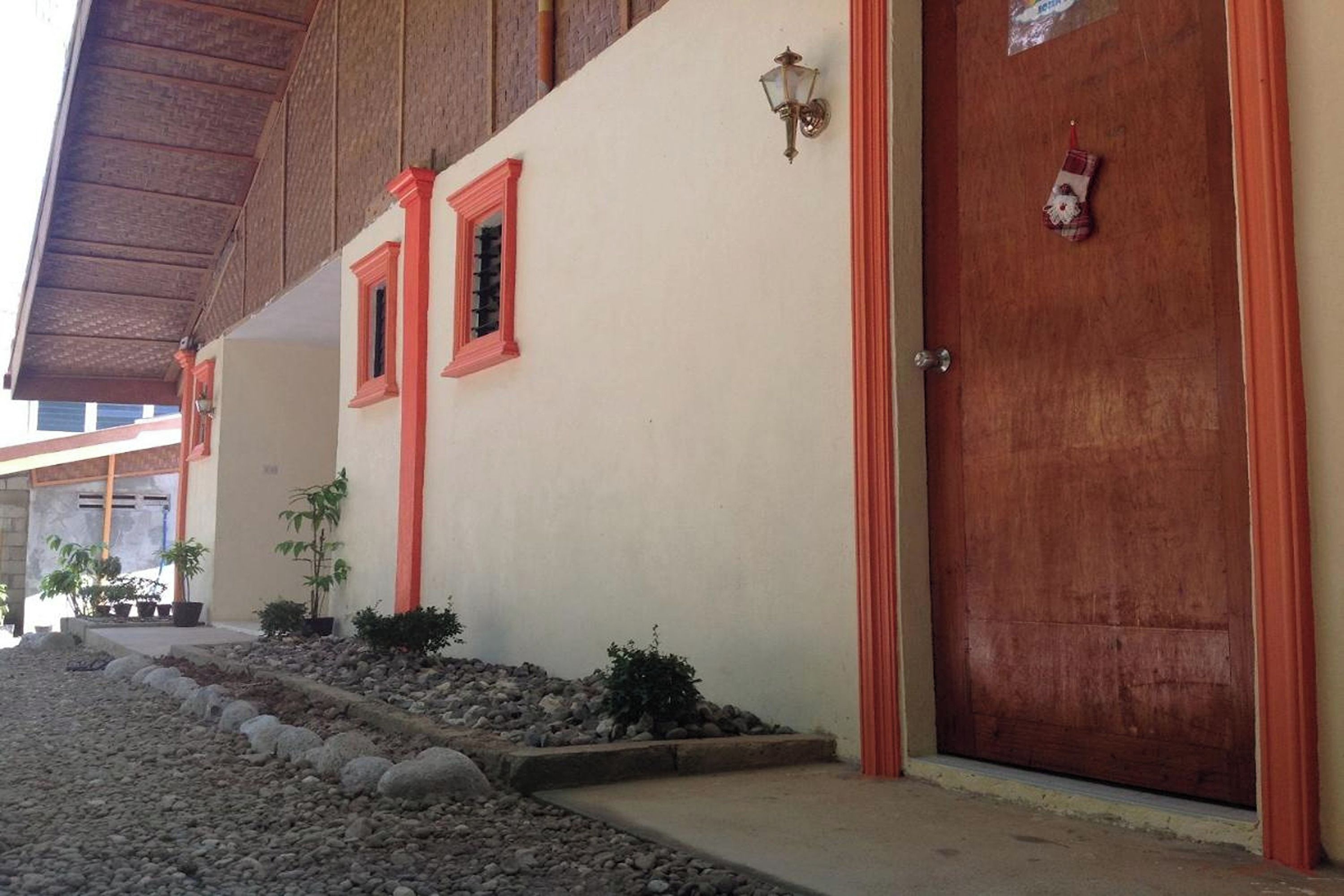 Un edificio con fachada color crema, ventanas de marco rojo, una puerta de madera y un pequeño jardín con rocas y plantas.