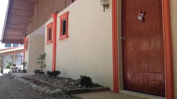 Un edificio con fachada color crema, ventanas de marco rojo, una puerta de madera y un pequeño jardín con rocas y plantas.