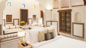 Una camera d'albergo lussuosa con un letto grande, una poltrona e un tavolino con una lampada. Sono presenti elementi decorativi e un'ampia porta in legno.