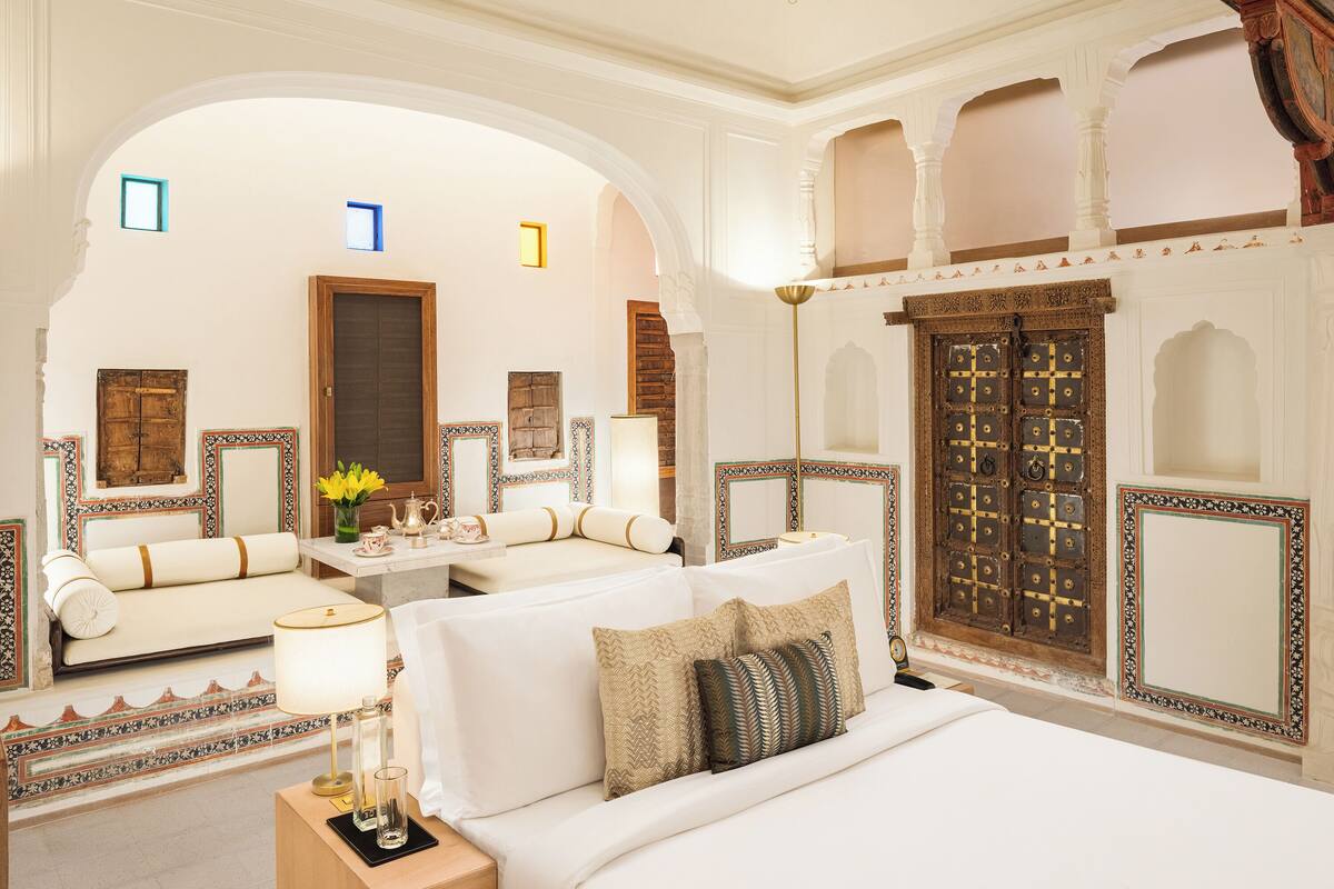 A luxurious hotel room with a large bed, a sofa, and a small table with a lamp. There are decorative elements and a large wooden door.