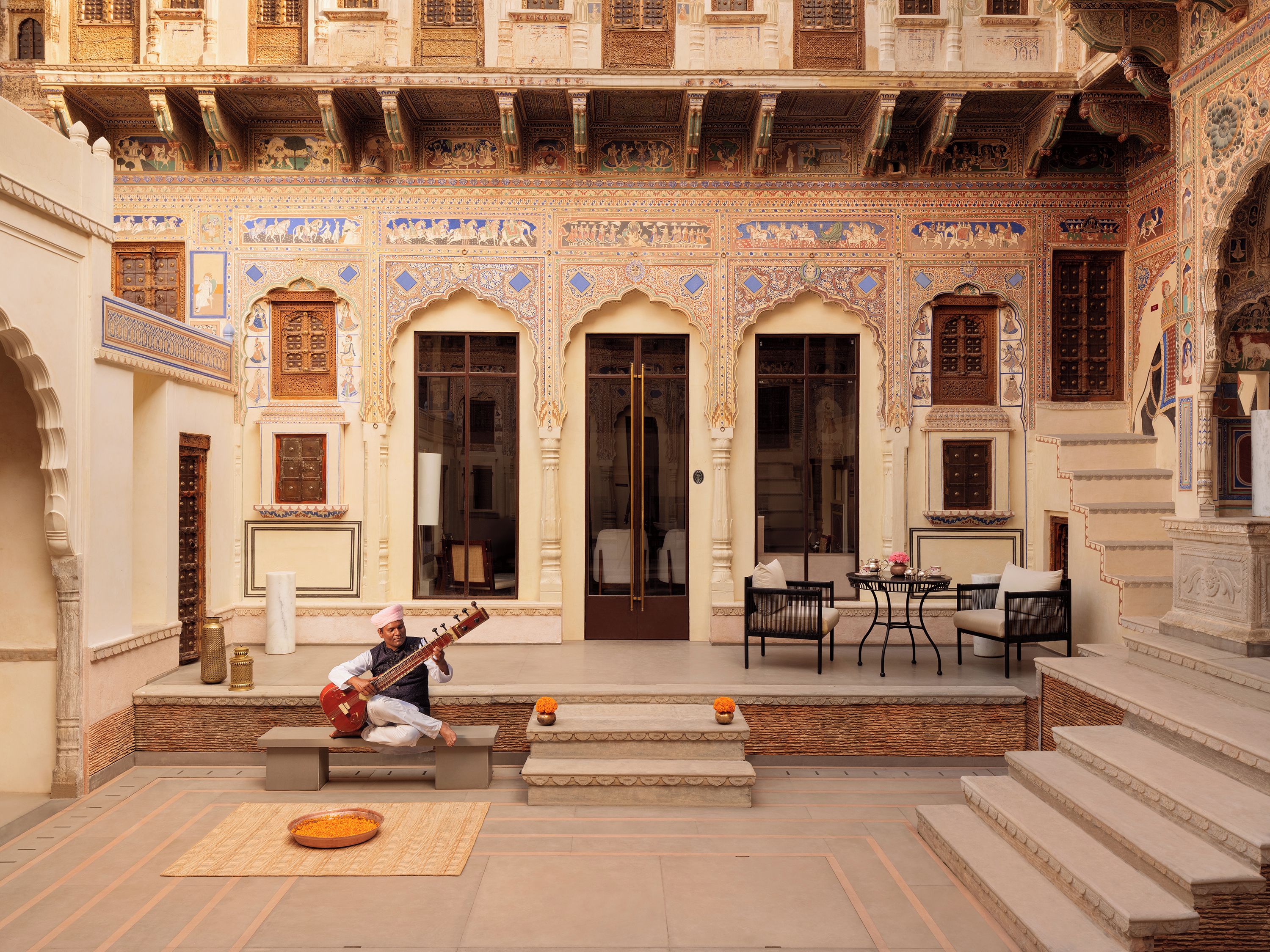 A courtyard with intricate architectural details, a musician playing a string instrument, and a seating area with chairs and a table.