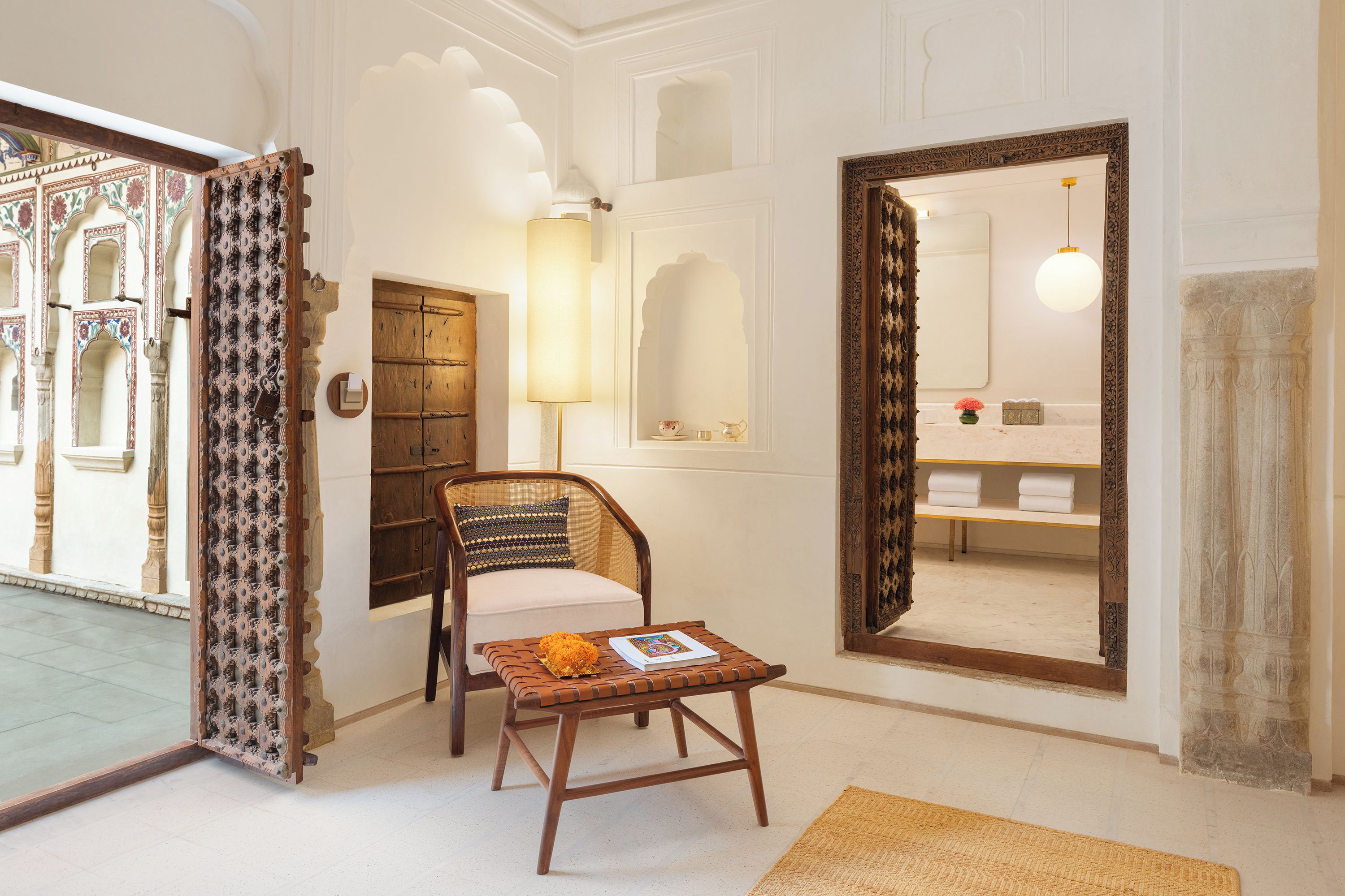 A room with a wooden chair, a small table with a book, a large mirror, and a door leading to a bathroom with a towel rack.