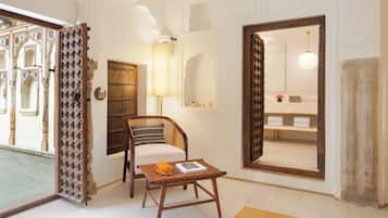 A room with a wooden chair, a small table with a book, a large mirror, and a door leading to a bathroom with a towel rack.