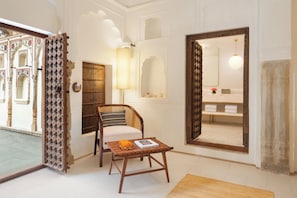 A room with a wooden chair, a small table with a book, a large mirror, and a door leading to a bathroom with a towel rack. - Gulab Haveli - Ihcl Seleqtions, Mandawa (Jhunjhunu)