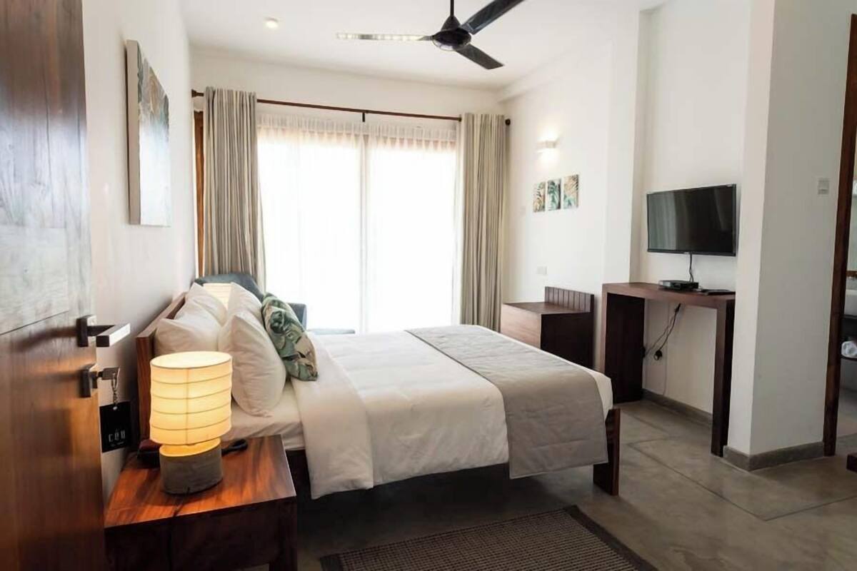 A modern hotel room with a bed, bedside table, lamp, TV, and curtains.