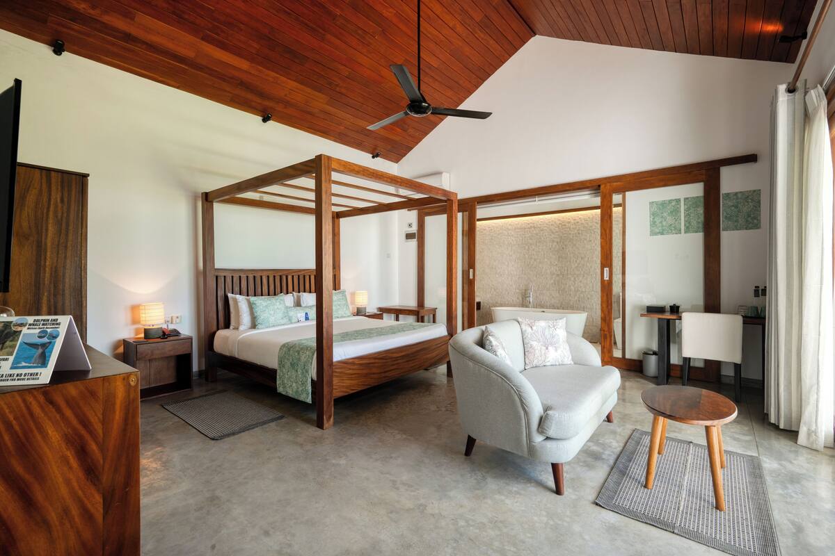 A modern bedroom with a four-poster bed, a sitting area with a sofa and armchair, and a small wooden table.