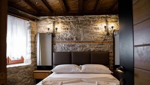 A room with a stone wall, a wooden ceiling, a bed with white linens, two bedside lamps, and a nightstand with a lamp. - Furra Hotel (Gjirokastër)
