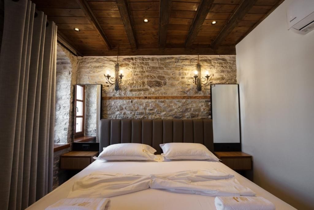 A hotel room with a stone wall, wooden ceiling beams, a large bed with white linens, two bedside tables, and wall-mounted sconces.