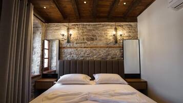 A hotel room with a stone wall, wooden ceiling beams, a large bed with white linens, two bedside tables, and wall-mounted sconces.