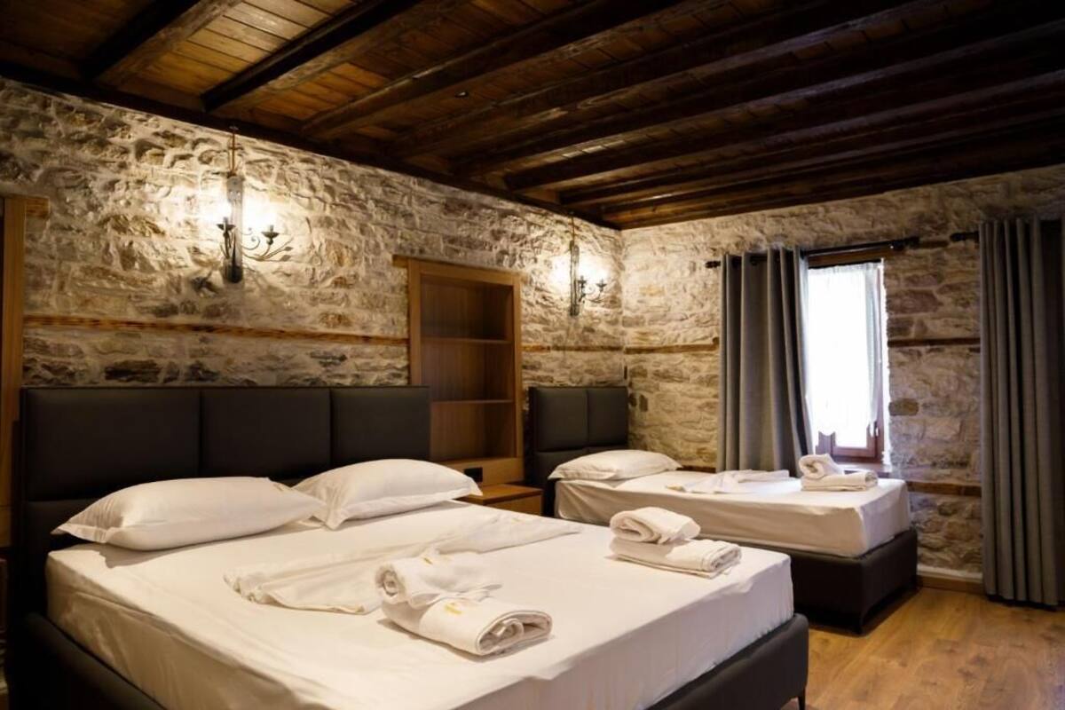 A room with two beds, stone walls, wooden beams, and a window with curtains.