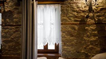 A stone-walled room with a bed, a window with curtains, and a wall-mounted light fixture.