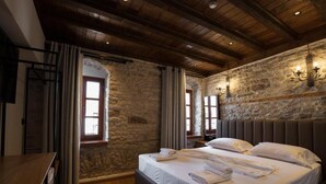 A bedroom with a stone wall, wooden ceiling beams, a large bed with white linens, and a nightstand. - Furra Hotel (Gjirokastër)