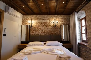 A hotel room with a stone wall, a wooden ceiling, a bed with white linens, and two wall-mounted lights. - Furra Hotel (Gjirokastër)