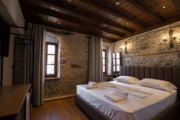 A bedroom with a stone wall, wooden ceiling beams, a large bed with white linens, and a nightstand. - Furra Hotel (Gjirokastër)