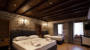 A bedroom with a stone wall, wooden ceiling beams, a large bed, two bedside tables, a TV, and a window with curtains. - Furra Hotel (Gjirokastër)