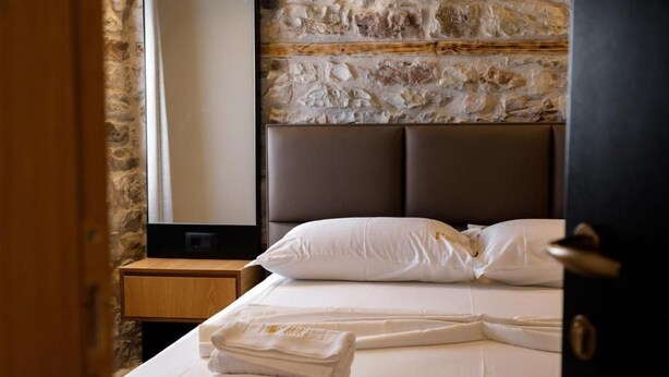 A hotel room with a stone wall, a bed with white linens, a wooden nightstand, and a folded towel on the bed. - Furra Hotel (Gjirokastër)