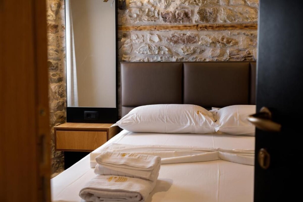 A hotel room with a stone wall, a bed with white linens, a wooden nightstand, and a folded towel on the bed.