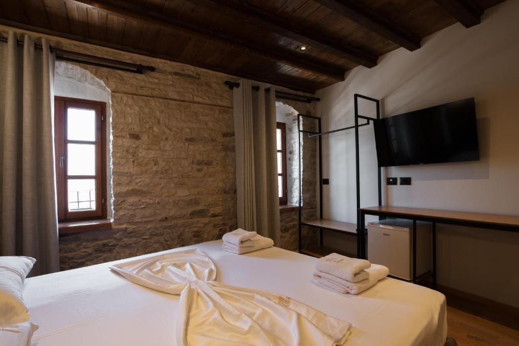 A hotel room with stone walls, a wooden ceiling, a bed with white linens, a TV mounted on the wall, and a small desk with a chair.