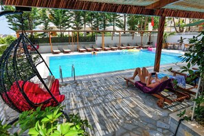 Zona de piscina con sillones de descanso, una silla colgante y un área de estar techada.