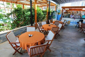 An outdoor patio area with round tables and chairs, a bar area with a bartender, and a view of greenery.