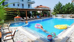 A swimming pool with lounge chairs, a covered seating area, and a building in the background.