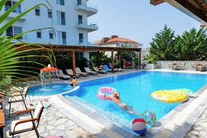 Una piscina con sillas de descanso, un área de estar techada y un edificio al fondo.