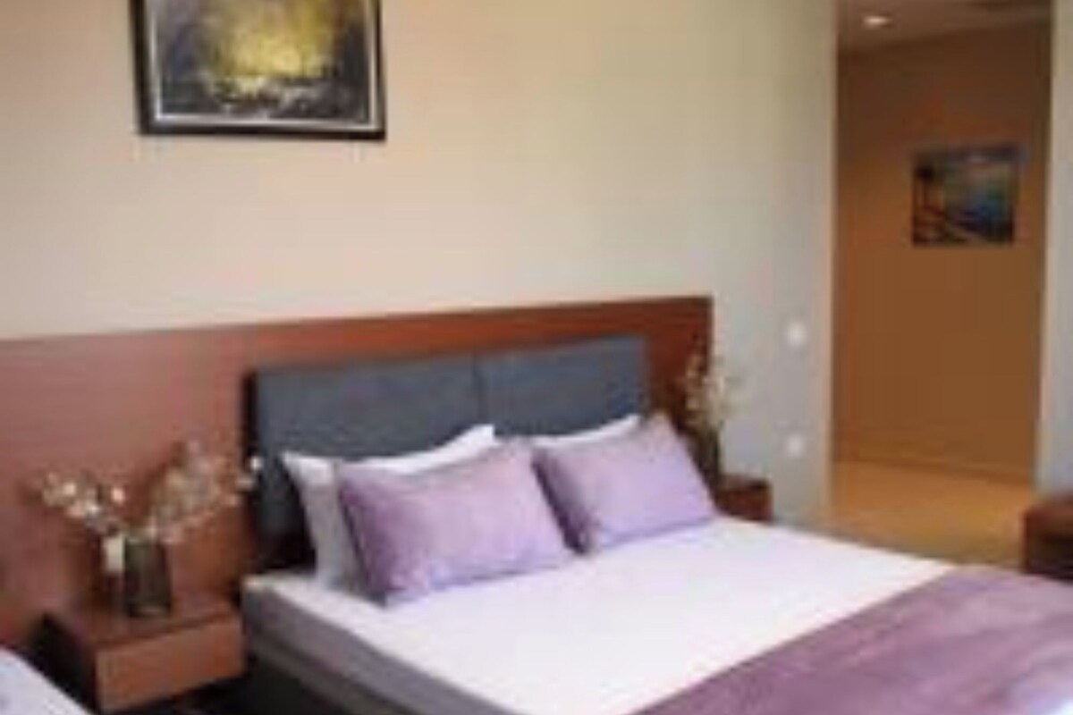 A hotel room with a large bed, a nightstand, a vase with flowers, and a painting on the wall.