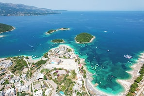 An aerial view of a coastal area with multiple islands, clear blue waters, and a developed beachfront with buildings and parked cars. - 6 Miles Hotel (Ksamil)