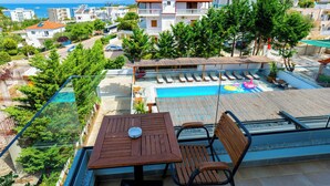 A balcony with a table and chairs overlooking a swimming pool and a residential area with buildings and trees.