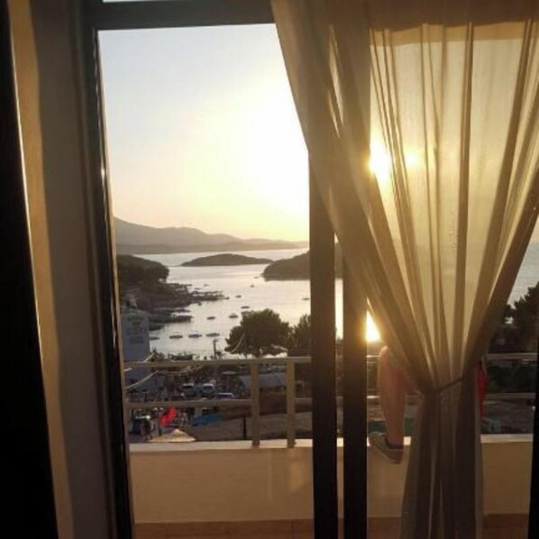 A view through a window with sheer curtains, revealing a coastal landscape with boats and buildings.