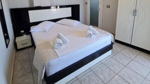 Un lit bien fait, avec du linge de lit et des serviettes blanches, une table de chevet, une armoire et un luminaire fixé au mur.