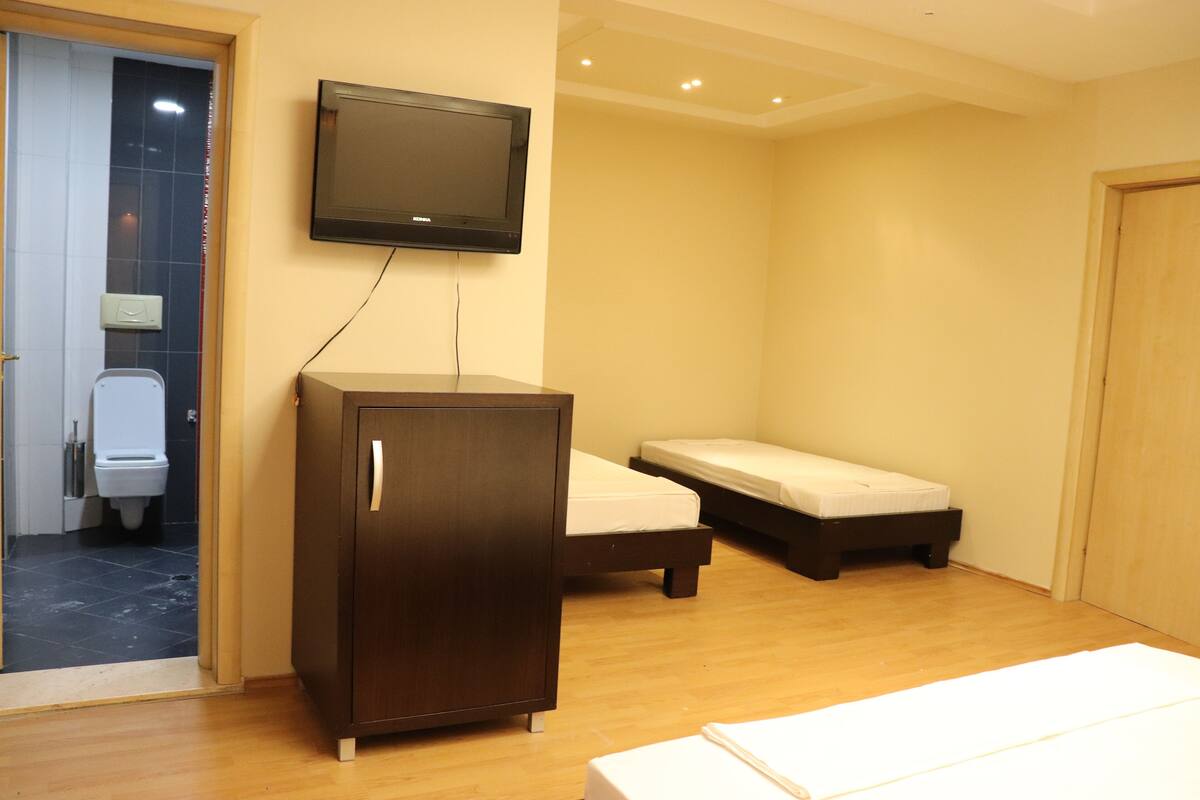 A room with a wooden cabinet, a bed with a mattress, a television mounted on the wall, and a toilet visible through an open door.