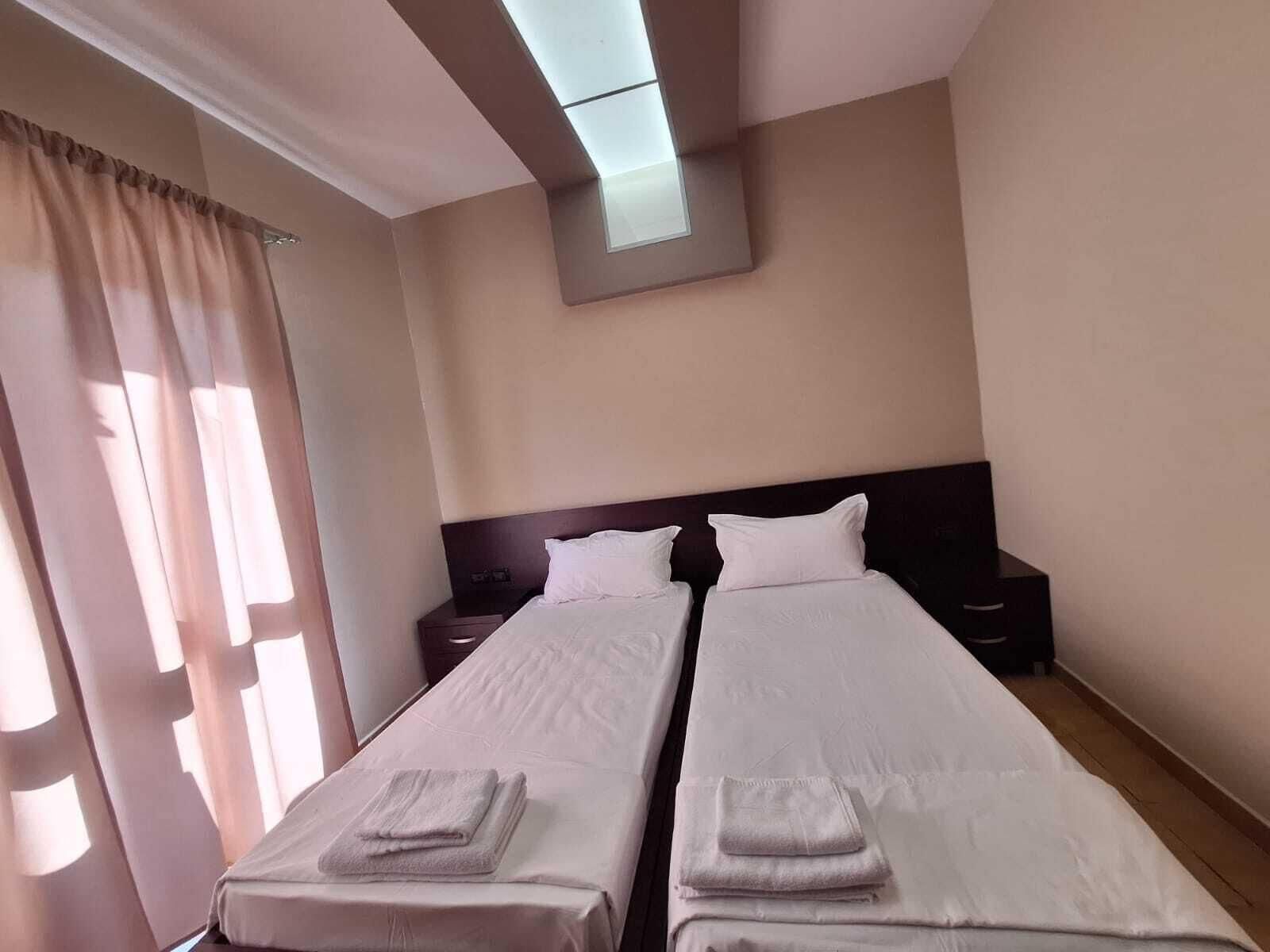 Une chambre d’hôtel avec deux lits simples, une table de chevet, un téléviseur fixé au mur et une fenêtre avec des rideaux.