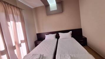 Camera d'albergo con due letti singoli, un comodino, TV a parete e finestra con tende.