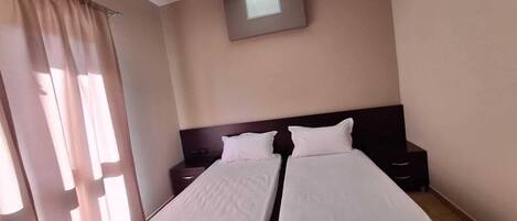 A hotel room with two single beds, a nightstand, a wall-mounted TV, and a window with curtains.