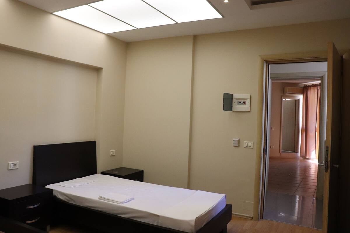 A bedroom with a bed, bedside tables, a ceiling light, and a door leading to another room.