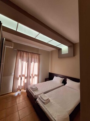 A hotel room with two beds, a ceiling light, a window with curtains, and a wall-mounted air conditioning unit. - Danis Hotel (Tirana)