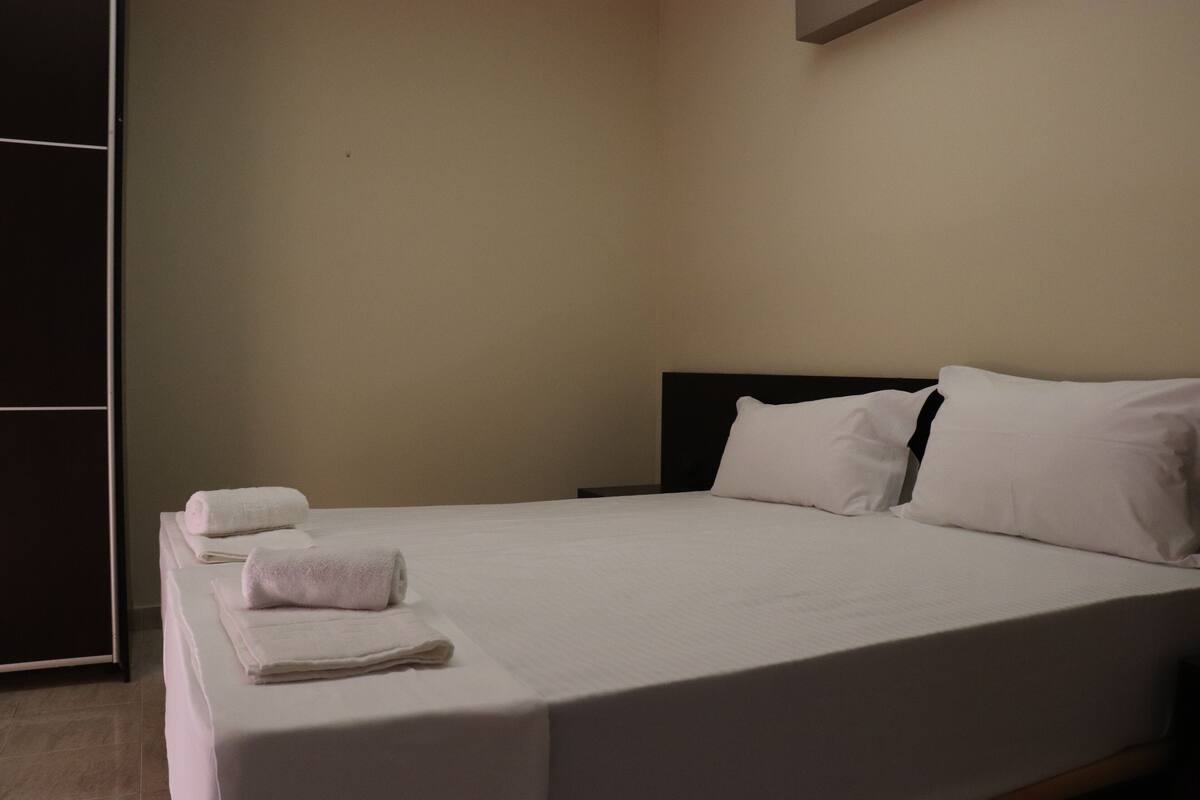 A neatly made bed with white linens and pillows, a dark headboard, and a plain wall.