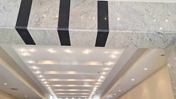 Eine Hotelhalle mit modernem Design, ausgestattet mit einer zentralen Treppe, einem Empfangsbereich und einer Decke mit eingelassener Beleuchtung und geometrischen Mustern.