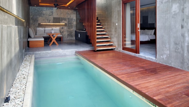 A modern outdoor pool area with a wooden deck, a seating area with cushions, and a staircase leading to an upper level. - Aqilah Villa Bali (Legian)